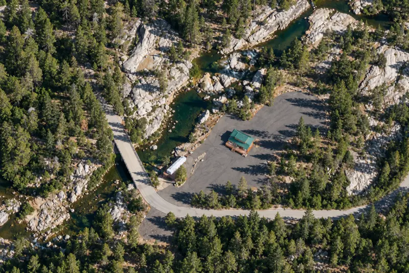 Aerial view: a cabin with a green roof on a paved clearing among pines; by the bridge over the river, the white sauna trailer with a deck and steps down to the pool