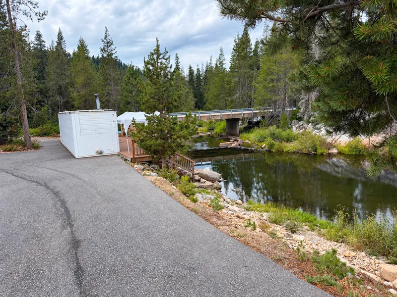 The sauna from outside: a white trailer with a chimney by the road, the deck, the river pool and a concrete bridge