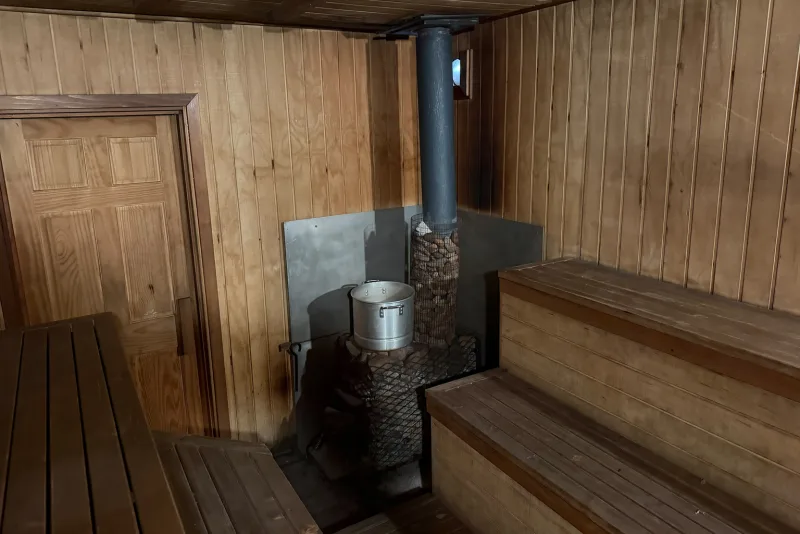 Inside the sauna: a wood-burning stove with a chimney pipe, a water tank on the stones, benches on two tiers